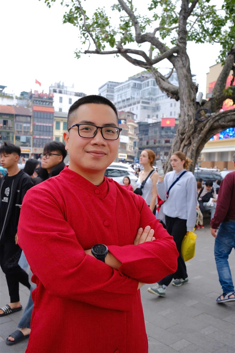 Chân dung anh Trần Trọng Trường mặc áo dài đỏ, khoanh tay cười tươi bên Hồ Gươm, Hà Nội